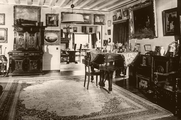 Een zwart-wit foto uit het verleden waarop een ander deel van de zitkamer te zien is met een vleugel en erg veel kunst aan de muren