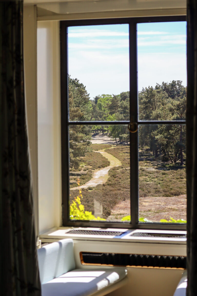 Een raampartij met uitzicht op bos en een wandelpad